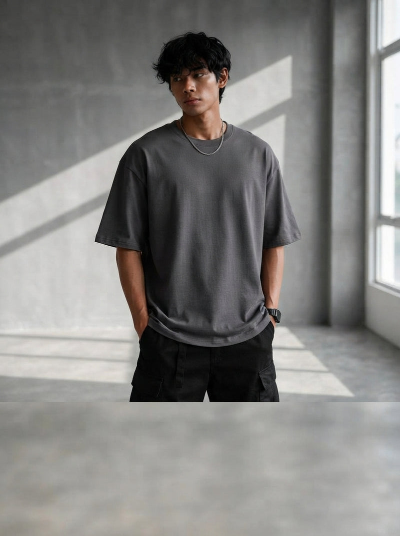 A South Asian man wearing an oversized charcoal t-shirt in a minimalist, sunlit studio setting.